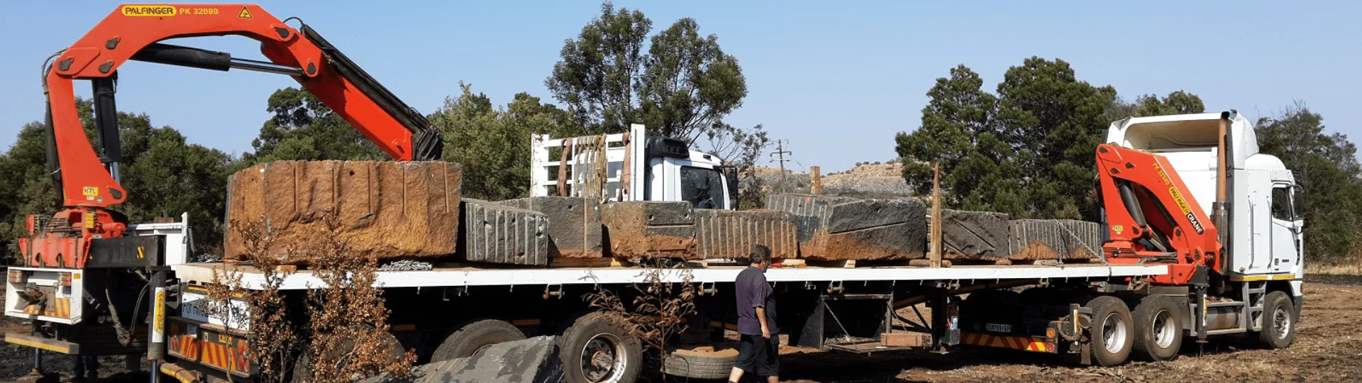 A large flatbed truck is parked in a field. It's loaded with bales of hay and caged equipment. Two cranes with 