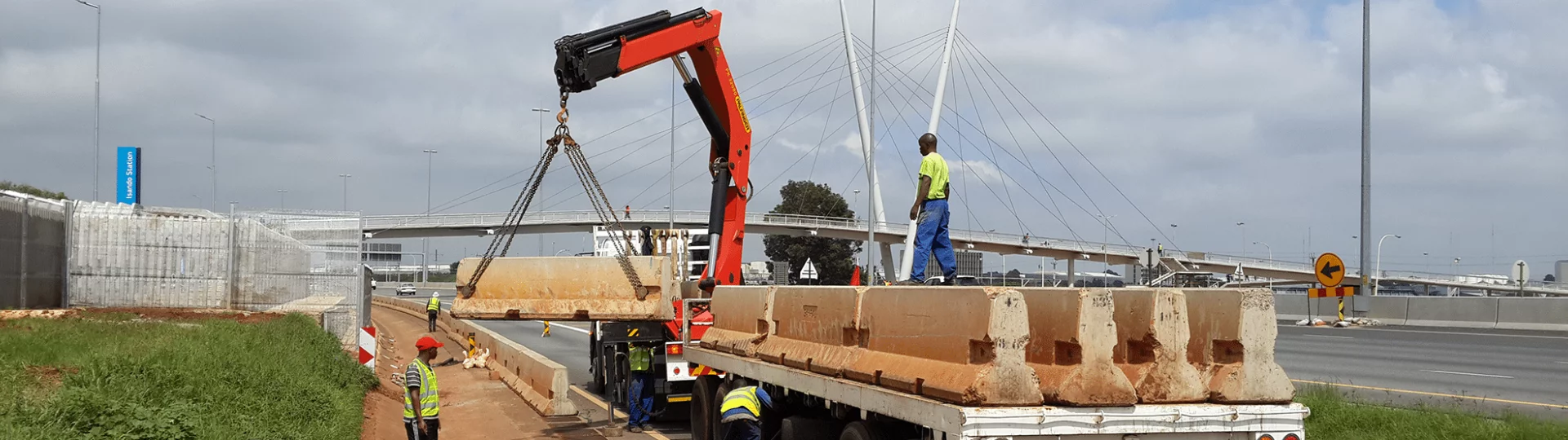 Truck with a crane unloading large concrete blocks on the roadside near a highway. Workers in safety vests guide the process. Overhead, a cable-stayed bridge is visible.